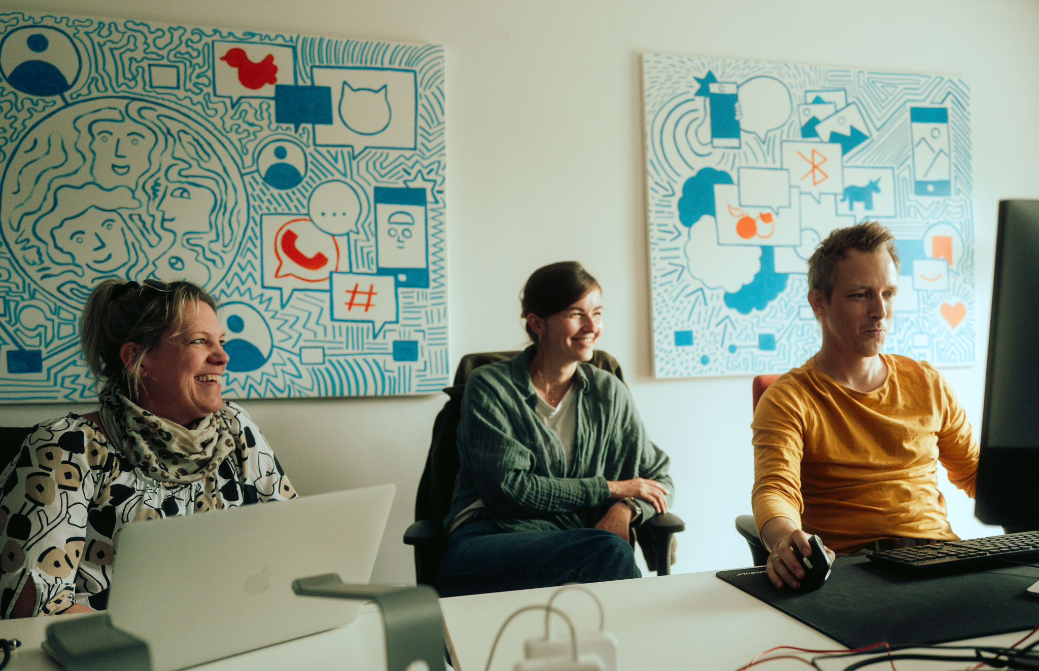 Drei Personen sitzen in einem Büro vor Computern und lächeln. Im Hintergrund hängen zwei farbenfrohe, grafische Kunstwerke an der Wand.