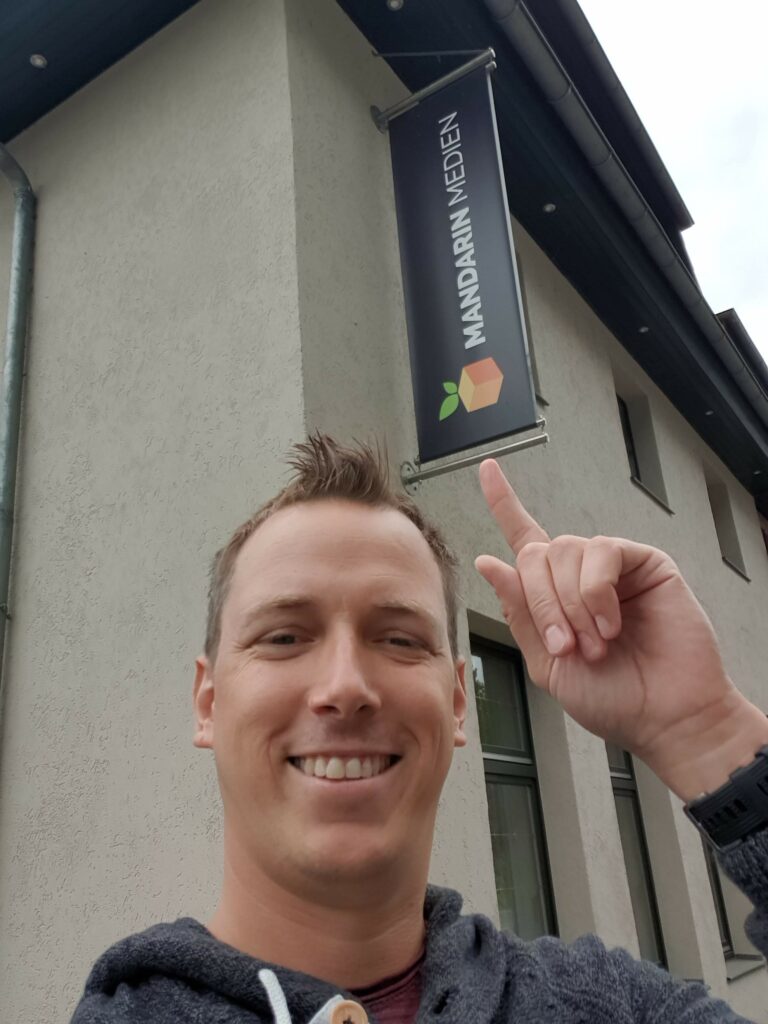 Marcel Rudolph, steht lächelnder vor einem Gebäude und zeigt mit dem Finger auf ein Schild, auf dem "Mandarin Medien" und ein oranges Würfel-Mandarin-Logo mit grünem Blatt zu sehen sind.