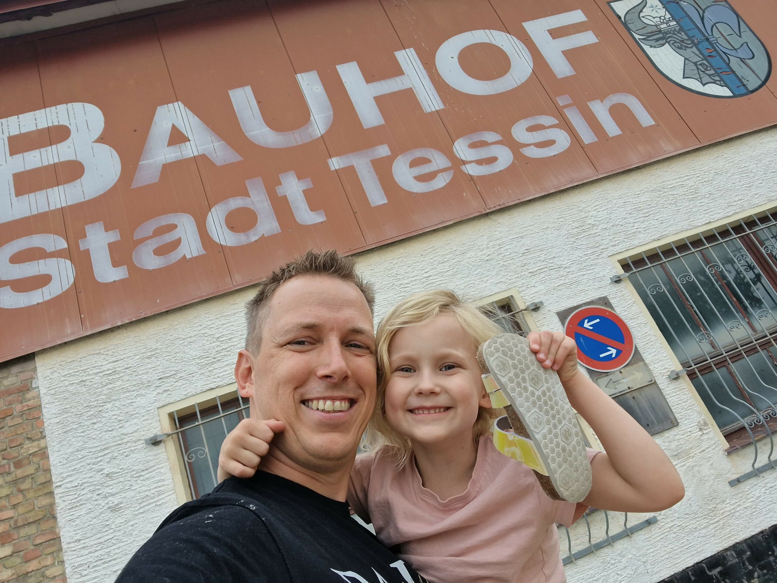 Mann und Kind posieren lächelnd für ein Selfie vor dem Gebäude des Bauhofs der Stadt Tessin. Im Hintergrund ist eine große Beschriftung "BAUHOF Stadt Tessin" auf einem rostbraunen Schild sowie ein Stadtwappen und ein Verkehrsschild zu sehen. Das Kind hält fröhlich einen Schuh hoch.